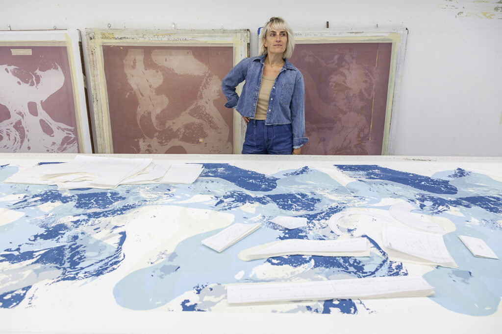 A photo of a blonde woman, the artist Aimee Koran, standing behind a table with a pattern of blue markings and assorted cut-out paper shapes laid overtop.