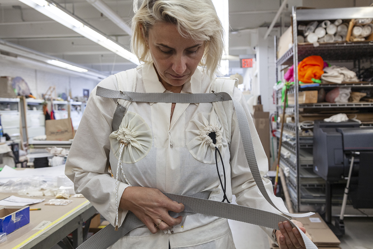 A photo of a white woman with blonde hair, the artist Aimee Koran, trying on a white jumpsuit with drawstrings positioned over her chest. She is stretching out a strap of fabric, presumably to tighten or measure it according to her body.