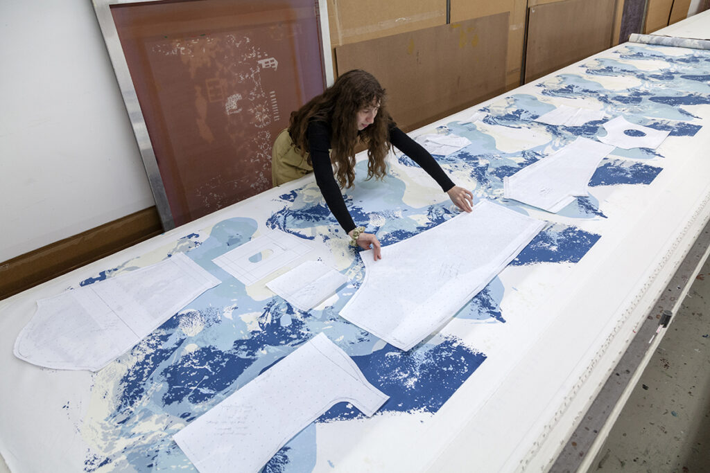 A photo of a brunette woman artist leaning over a table to spread out a cut-out paper shape overtop a long blue printed surface. There are similar white shapes cut out and laying nearby.