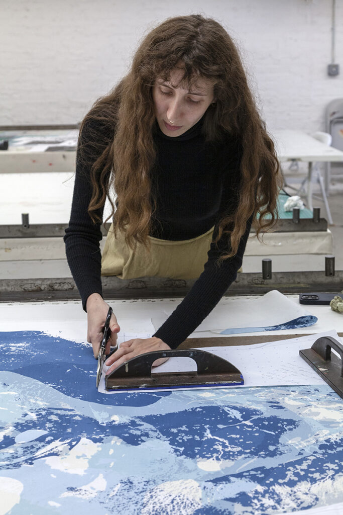 A brunette woman artist leans over a table to cut a piece of fabric with blue patterns printed on it. She is holding scissors with her right hand while pressing down on a weight with the other.