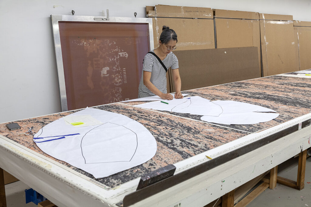 A photo of a woman artist working on a table with a large paper shape cut out and laid over a fabric pattern.