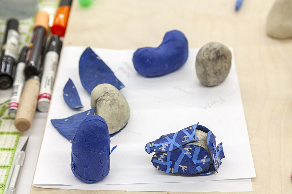 A photo of small rock-like forms covered in layers of bands of blue tape. One on the bottom-right appears to be deconstructed, exposing the many layers of tape and the form underneath.