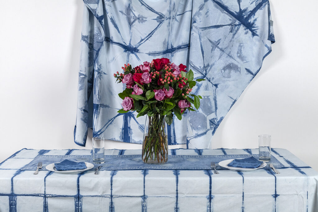 A vase of flowers sits atop a tablecloth printed with indigo, in front of an indigo-printed backdrop.