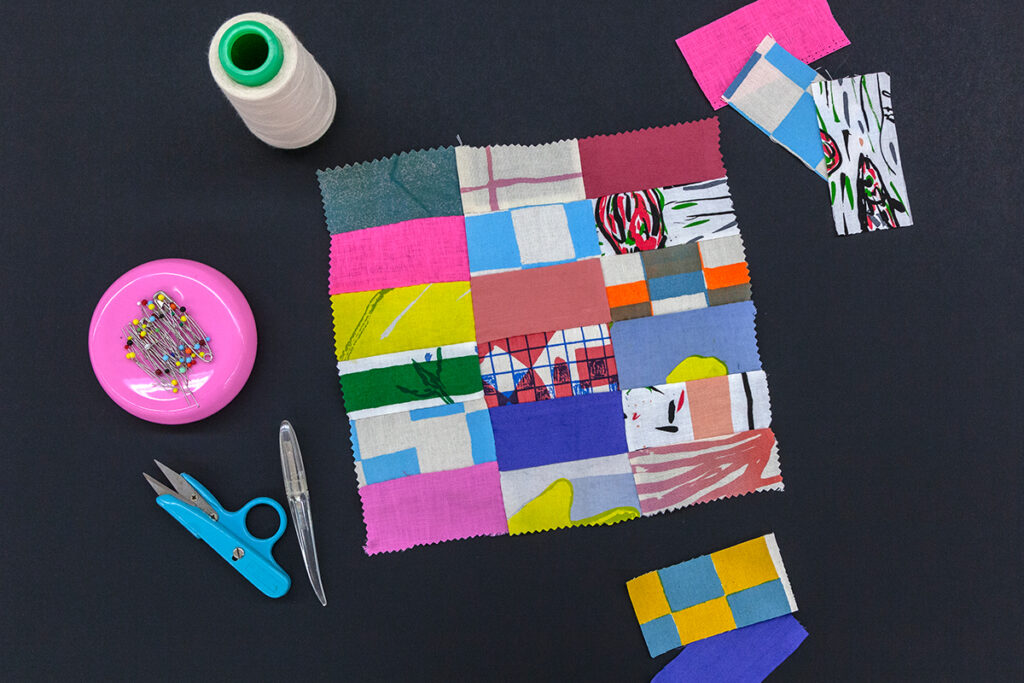 A sample of materials, including quilted fabric squares featuring disparate swatches of colors and patterns, a spool of thread, sewing needs, and other tools.