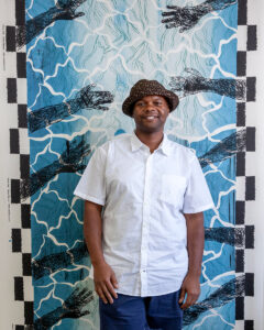 A portrait of Anthony Folks, a young Black man with a bucket hat and short-sleeve white button-down shirt. He is standing in front of a printed artwork with hands reaching inward toward a blue water-like environment.