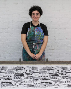 A portrait of the artist Hannah Moog. A white woman with dark curly hair. She is wearing an apron and standing in front of a large printed artwork laid on a table.