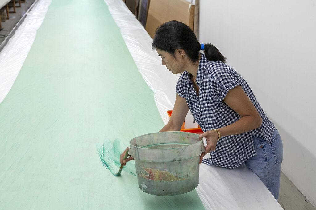 A photo of the artist Julia Chiang, an Asian American woman, painting with green ink on a long table. She is holding a bucket with her left hand while painting with her right hand.