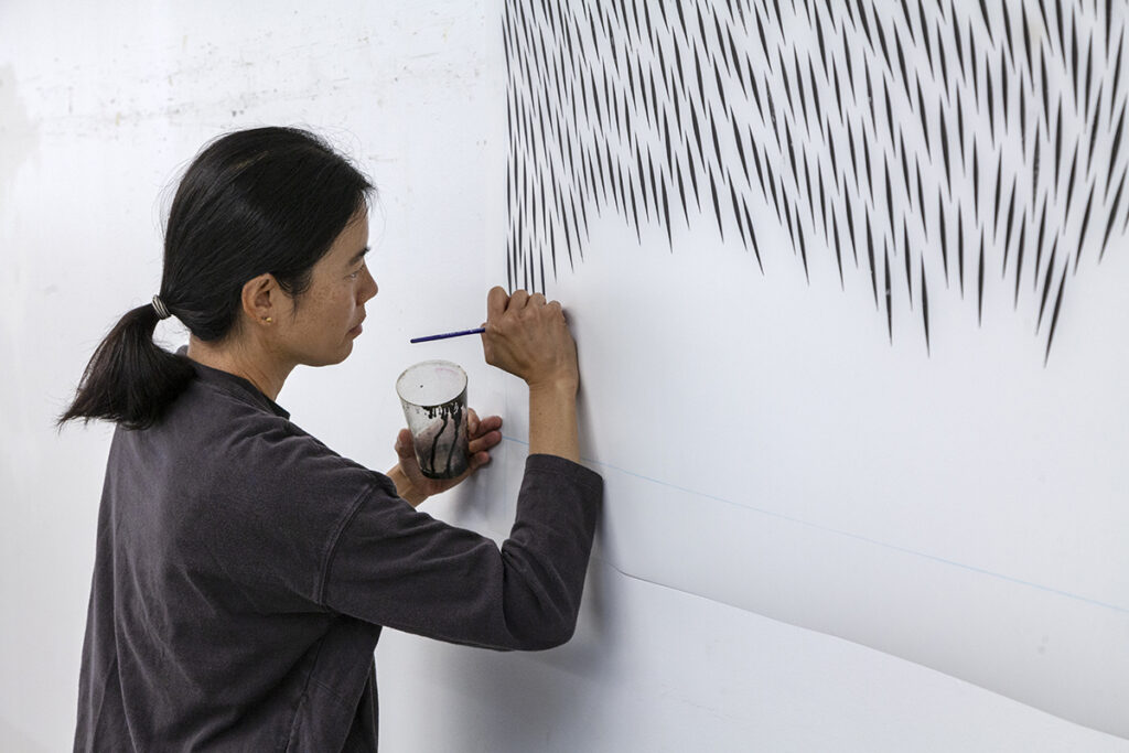 A photo of the artist Julia Chiang, an Asian-American woman, drawing abstract marks on a surface pinned to a wall. She is right handed and is holding a small cup of ink with her left hand.