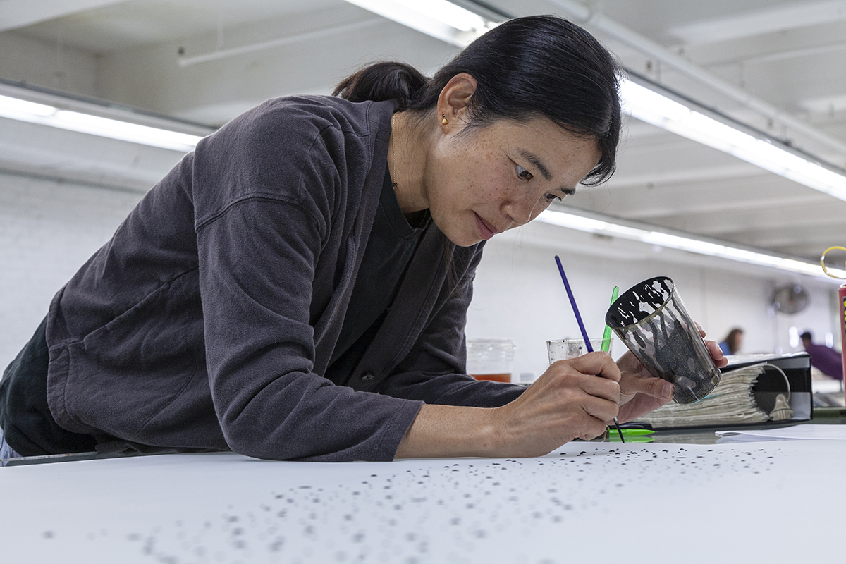 A photo of the artist Julia Chiang, an Asian-American woman, painting with a fine-tipped brush on a surface.