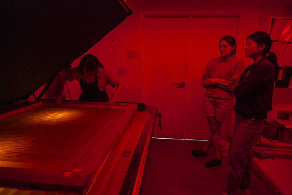 A photo of three people in a deeply red-lit room placing artwork on an exposure table.