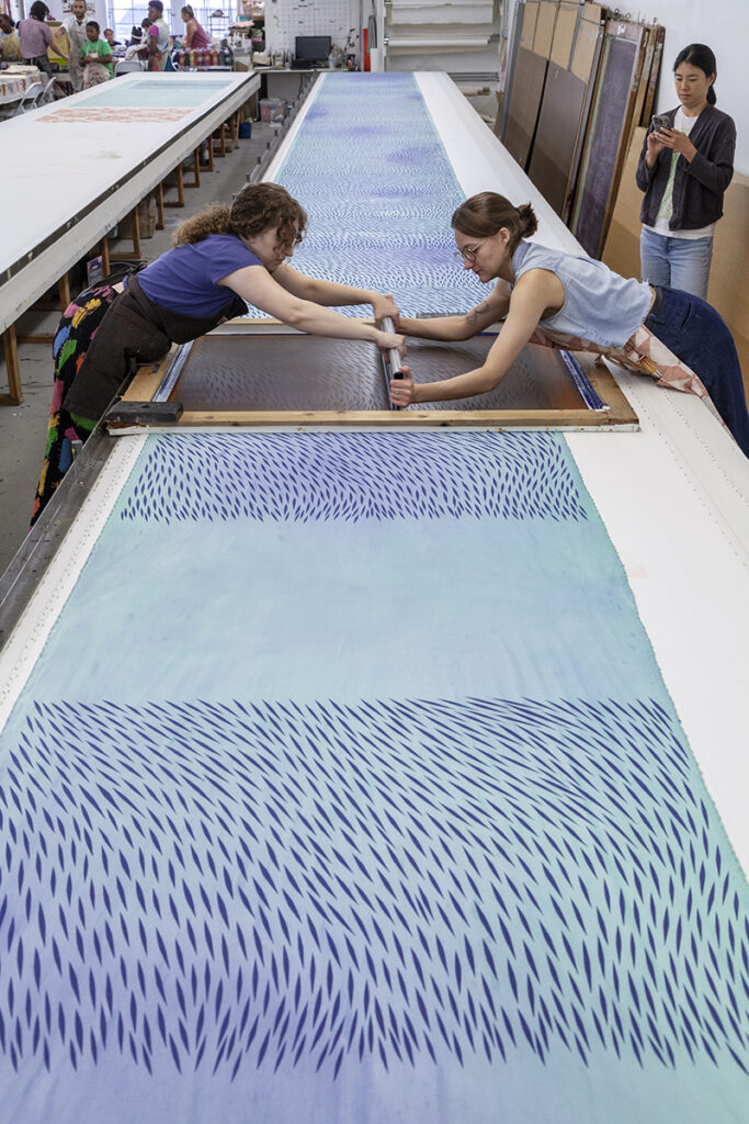 A photo of two artists screenprinting on a long table as another artist looks on. They are printing an abstract pattern over a sea blue ground.