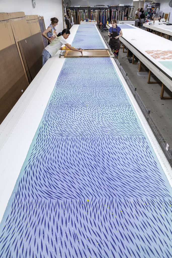 A photo of three people screenprinting on a very long table. They are printing an abstract pattern over a sea blue ground.