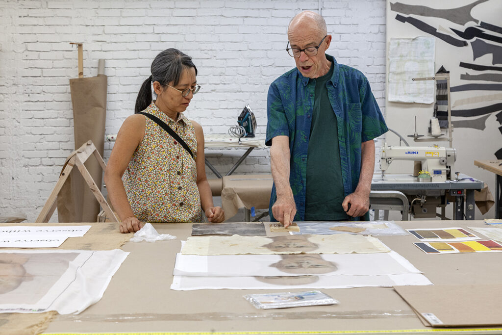 A photo of John Killacky, an older white man with glasses, points toward portrait studies on a table while Nami Yamamoto, a Japanese woman with glasses, looks on.