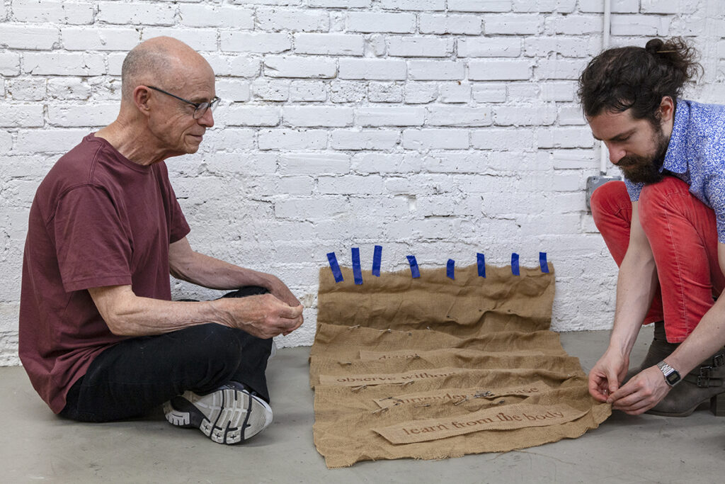 A photo of two white men squatting on the floor with an artwork made of burlap between them on the floor.