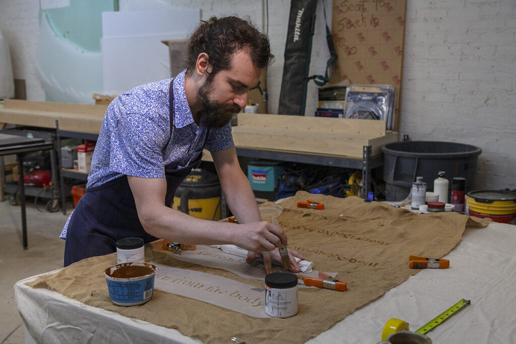 A photo of a bearded man with dark hair painting on a burlap piece of art on a table.