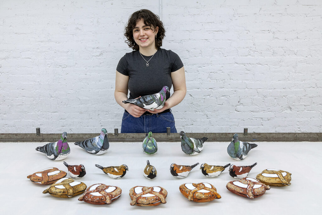 A young white woman, Oonagh McKenna, stands behind a table showcasing her artworks. She is holding a stuffed pigeon while the table presents more pigeons, as well as stuffed sparrows and pretzels.