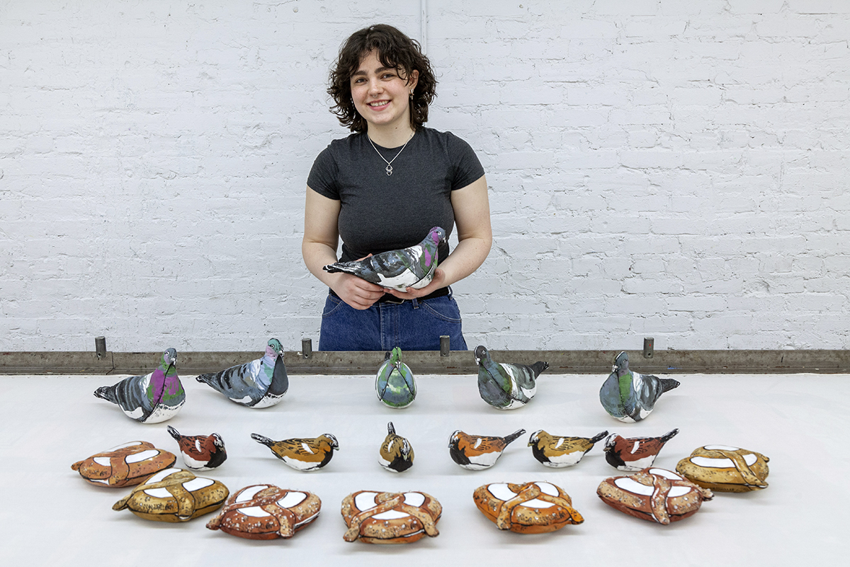 A young white woman, Oonagh McKenna, stands behind a table showcasing her artworks. She is holding a stuffed pigeon while the table presents more pigeons, as well as stuffed sparrows and pretzels.