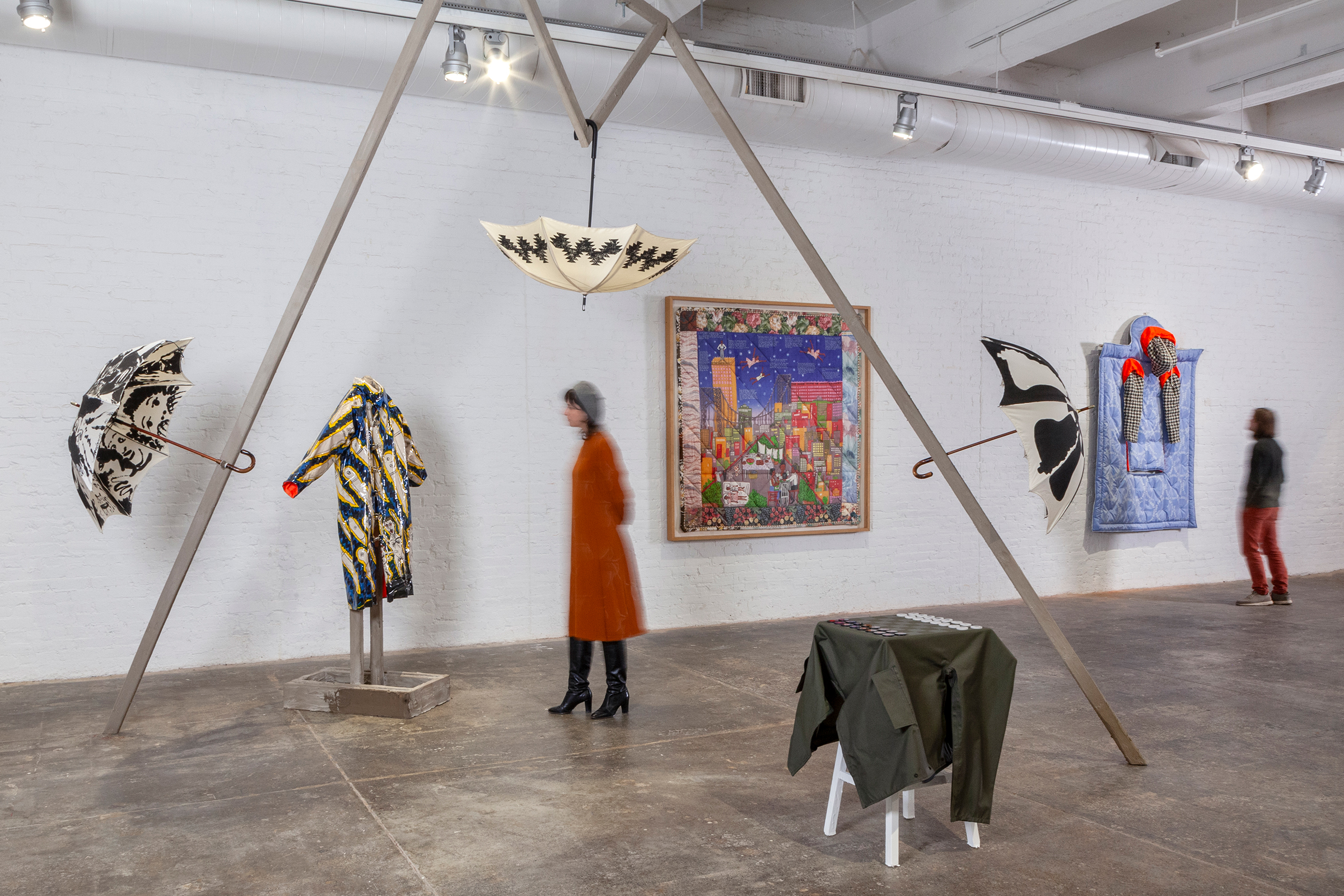 A gallery installation photo showing three umbrellas installed at various ends of a triangular armature with other garments and artworks installed on the floor or wall nearby.