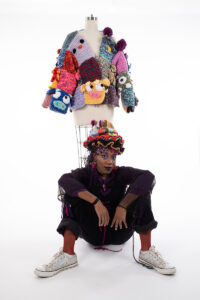 A Black artist, Kalila Jones, sits below a mannequin with arms resting across their knees. They are wearing a crochet hat with many layers of colors, a black and purple long sleeve garment, and white sneakers. On the mannequin, a colorful crochet garment features sculptural faces of cartoon-like characters.