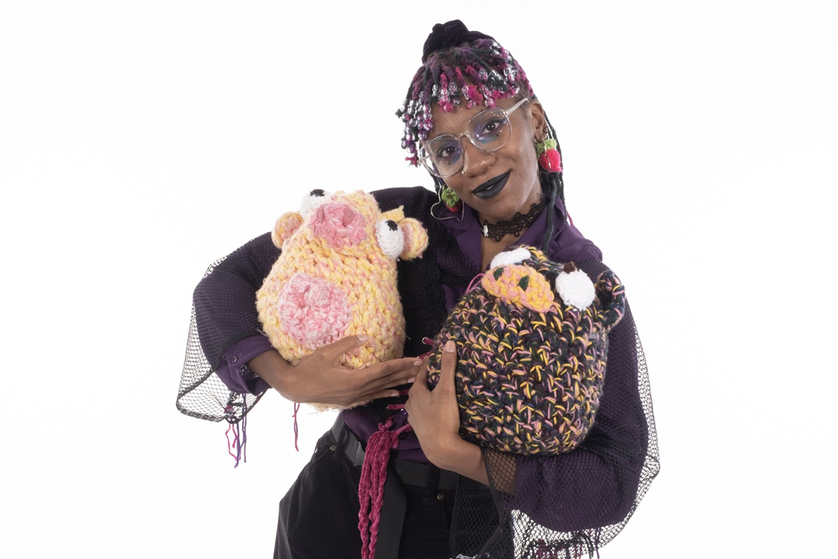 A Black artist, Kalila Jones, smiles while holding two cartoon-like animals made from crochet in their arms. Their hair is braided. They are wearing glasses and strawberry-like earrings, and a black and purple long sleeve garment.