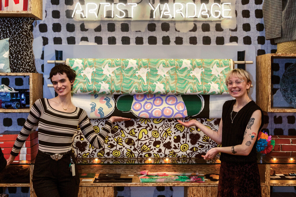 Two FWM Store Associates posing in front of the fabric yardage display in the FWM Store. There are three colorful, patterned fabrics hanging horizontally on dowels. Above the fabric there is a sign in neon light that reads "Artist Yardage."