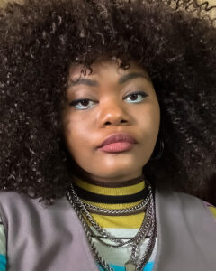 A portrait of Kierra Crafton, a Black woman with big curly hair. She is wearing a yellow turtle neck with necklaces and a gray shirt with a deep v-neck.