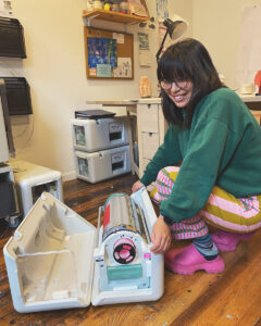 A young Chinese-American woman, Madeleine Conover, kneels in her studio to adjust a printer on her floor