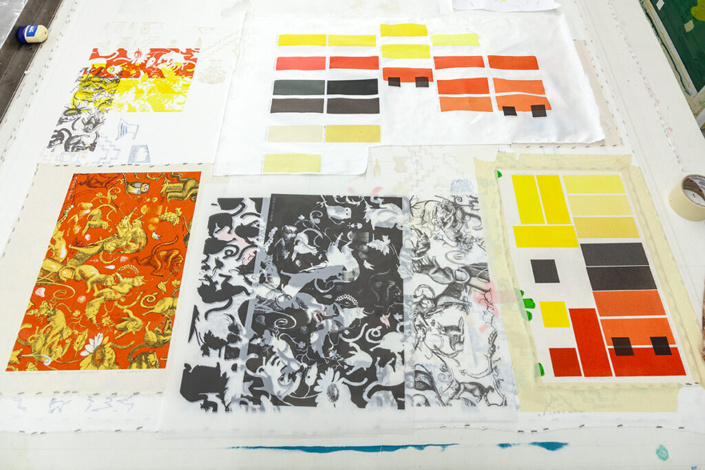 A photo of material and color tests. At the top and right are a number of bars of colors tinted in reds, oranges, yellows, and blacks. At the bottom-left is a pattern printed in those colors. To its right is the same pattern printed in black.