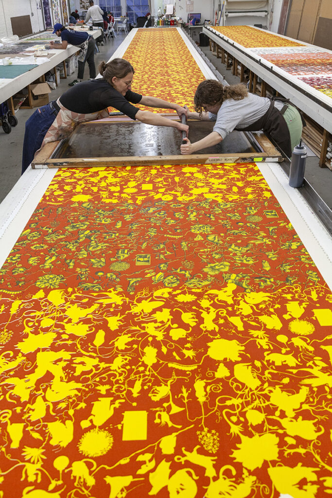 A photo of two women artists screenprinting a red/orange and yellow pattern on a long table.