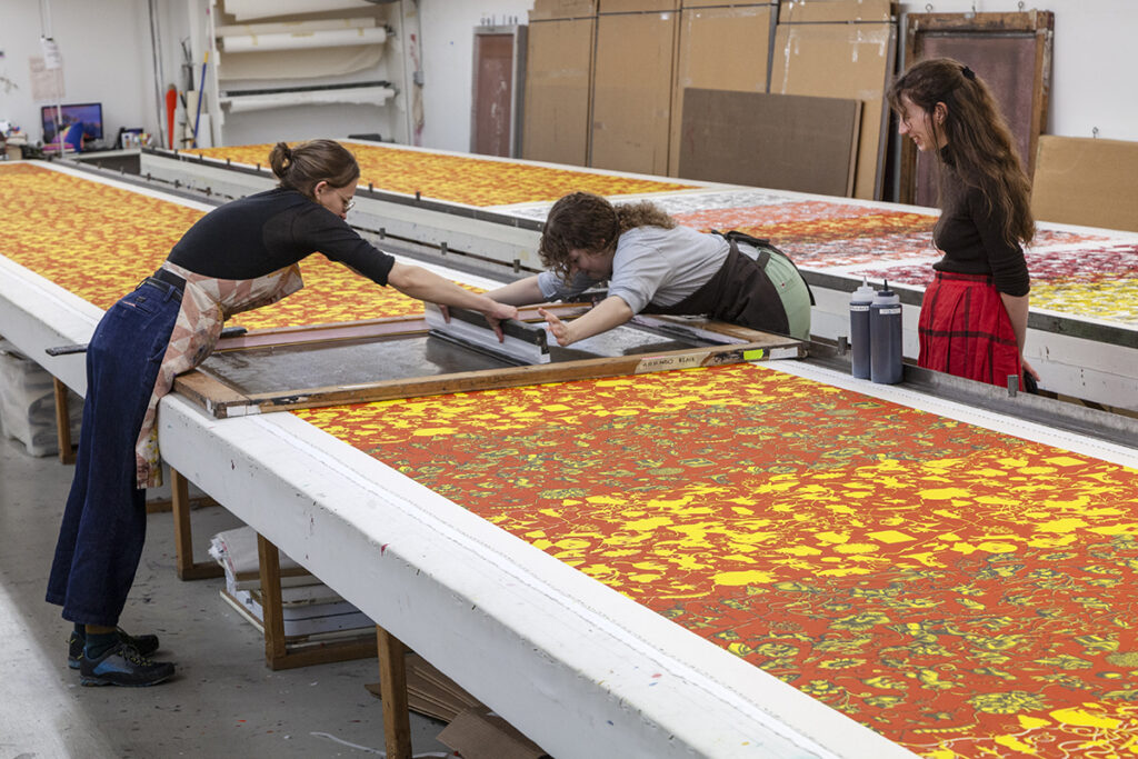 A photo of two women artists screenprinting a red/orange and yellow pattern on a long table. They are leaned over the table to push a squeegee along a silkscreen while a colleague looks on at the right of the image.