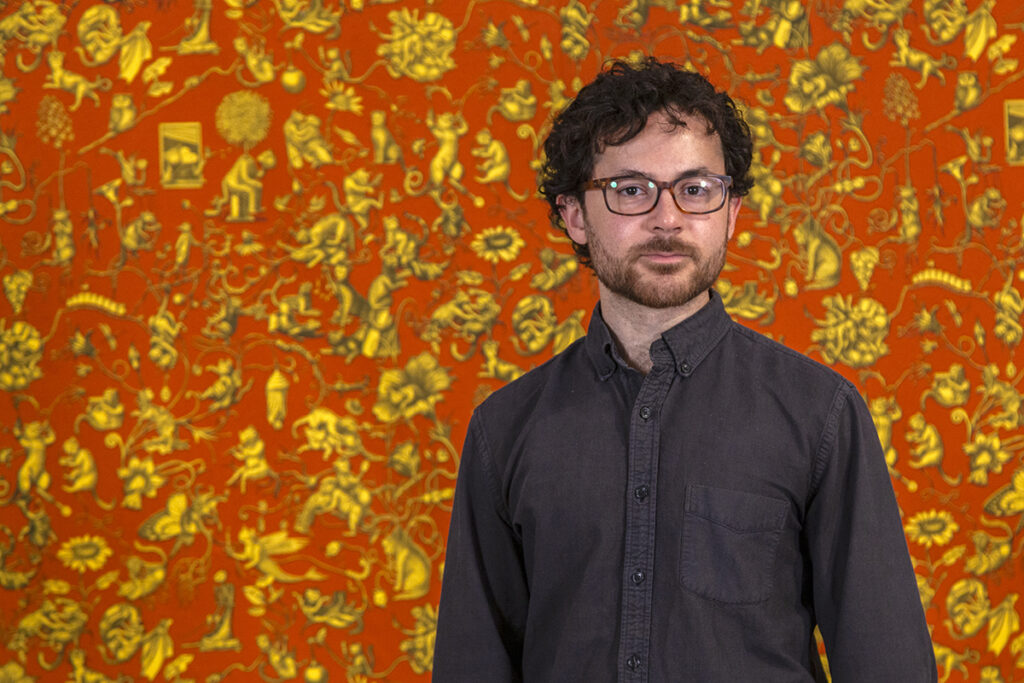 A portrait of Armando Veve, a white man with dark curly hair, a beard, and glasses, standing in front of his large tapestry. The artwork consists of many tiny illustrations in yellow with black outlines and details against an orange-red background.