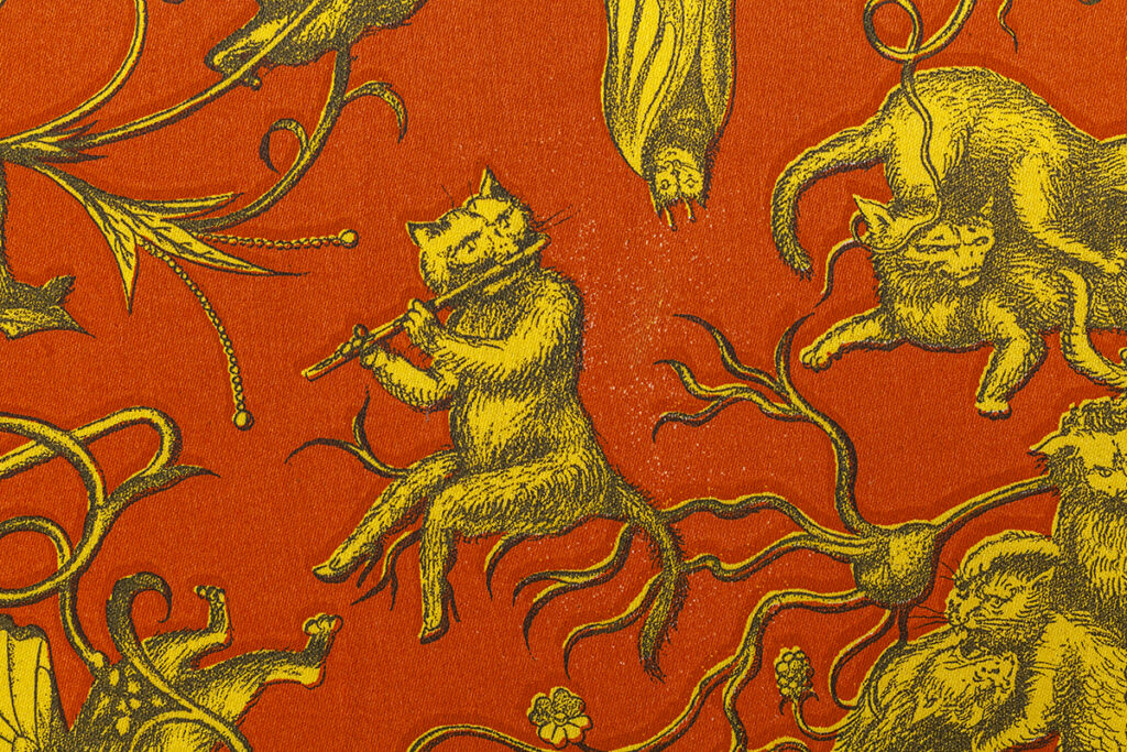 A detail of an illustrated pattern featuring a cat playing a flute while seated on a branch. Fighting cats and a bat hanging upside down appear near the periphery. The pattern is composed of three basic colors with a red/orange ground, yellow characters, and dark gray details.