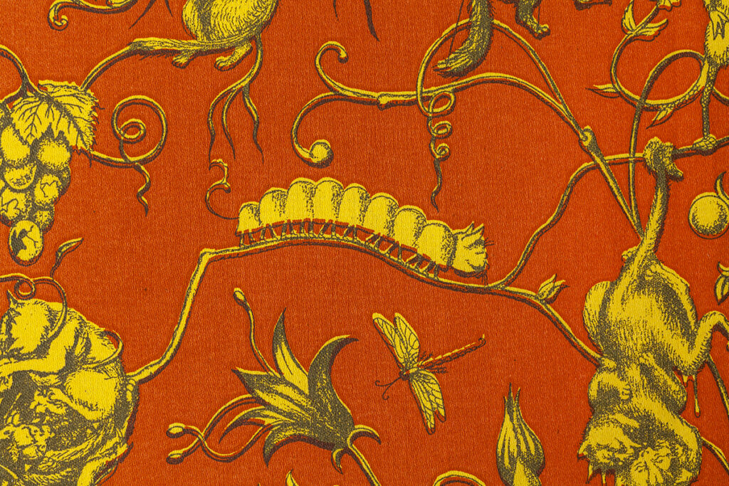 A detail of an illustrated pattern featuring a caterpillar inching along a thin vine with flowers, bugs, and spindly vines stretching about. The pattern is composed of three basic colors with a red/orange ground, yellow characters, and dark gray details.