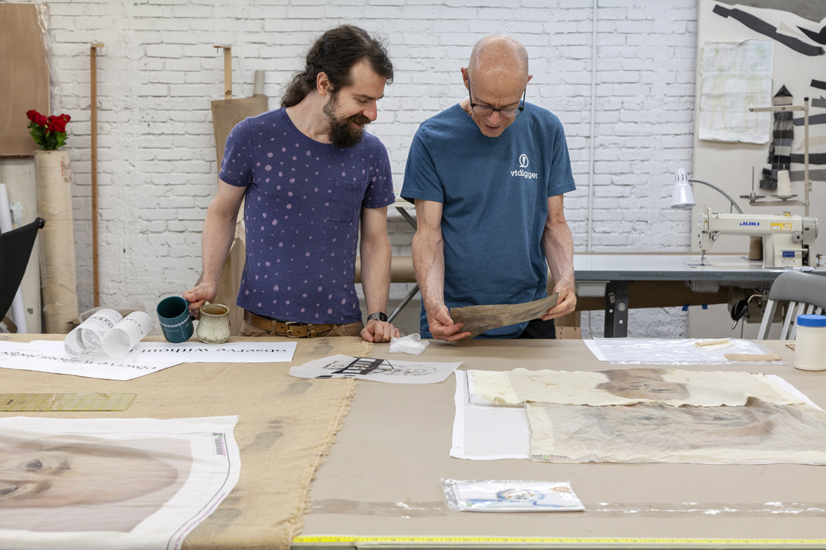 A photo of two white men working in an artist studio. The bald man on the right, John Killacky, shows a piece of material to the bearded man on the left, Allen West. On the table in front of them are more drawing samples.