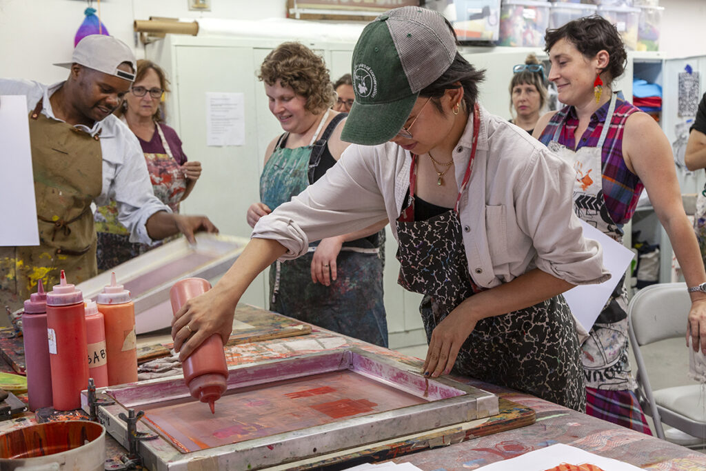 A young woman wearing a hat and apron squeezes ink from a bottle onto a silkscreen during a screenprinting class. People in the background are watching an instructor show off the results of a print.