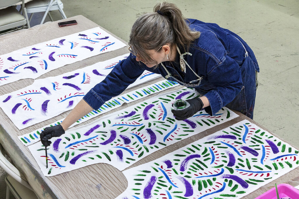 A woman standing over a table with pieces of a pattern to make a pair of pants. She is painting a vivid, fluid design on the fabric of the pants pattern.