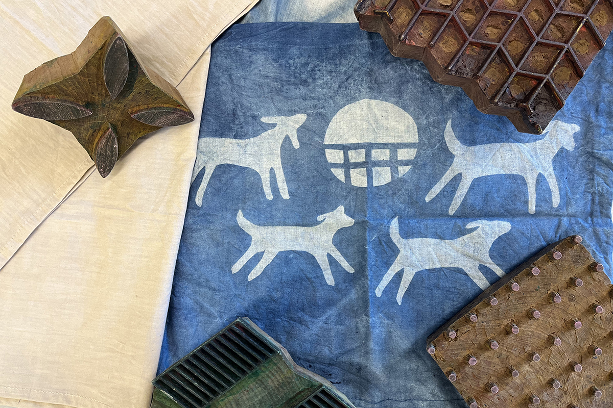 A photo of an indigo-dyed napkin with a motif of dogs. A variety of wooden blocks with different carved motifs are displayed around the napkin.