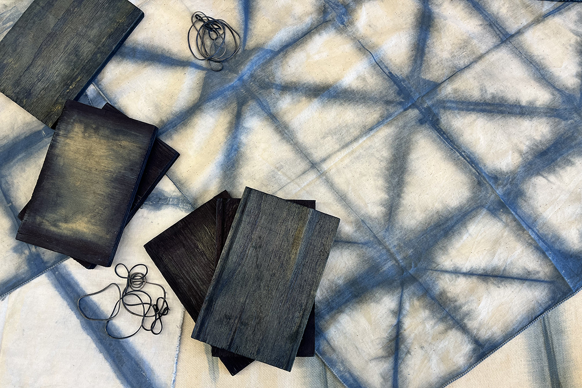 A photo of an indigo-dyed tablecloth with a triangular dye pattern. There are blue wooden panels of wood and rubber bands sitting on top of the fabric.