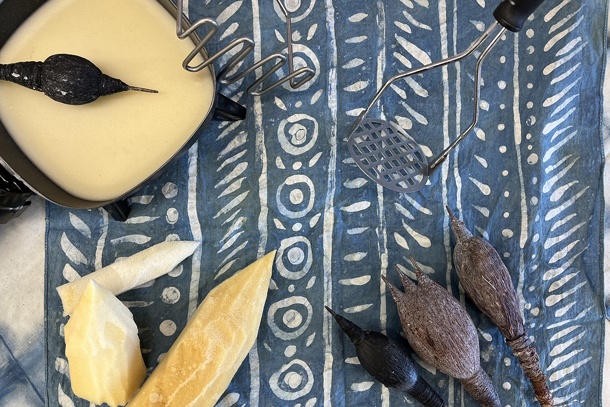 A photo of indigo-dyed fabric with white decorative pattern markings with a pot of wax and a variety of mark-making tools sitting on top of the fabric.