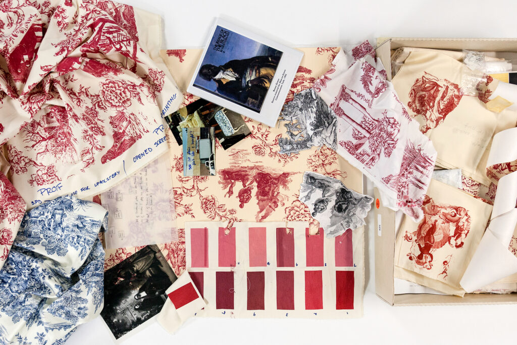 Archival contents spill out of a box including prints with red illustrations, photographs, and color swatches.