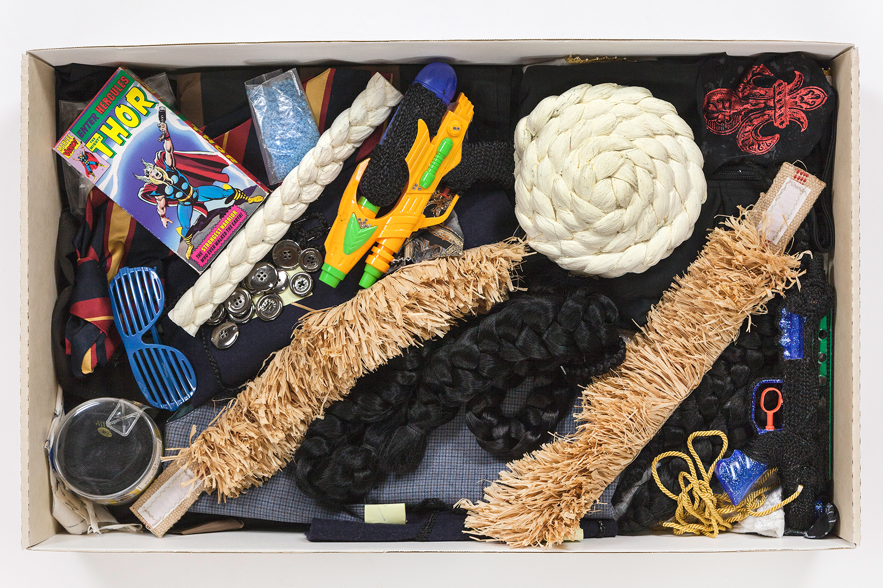 A rectangular box of material contents including woven yarn, raffia, photographs, a Thor VHS tape, plastic sunglasses, and a toy gun.