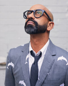 A portrait of the artist Brendan Fernandes, a brown-skinned man with a shaved head and black beard. He is looking up while wearing black-rimmed glasses, a blue-gray suit, and gray tie.