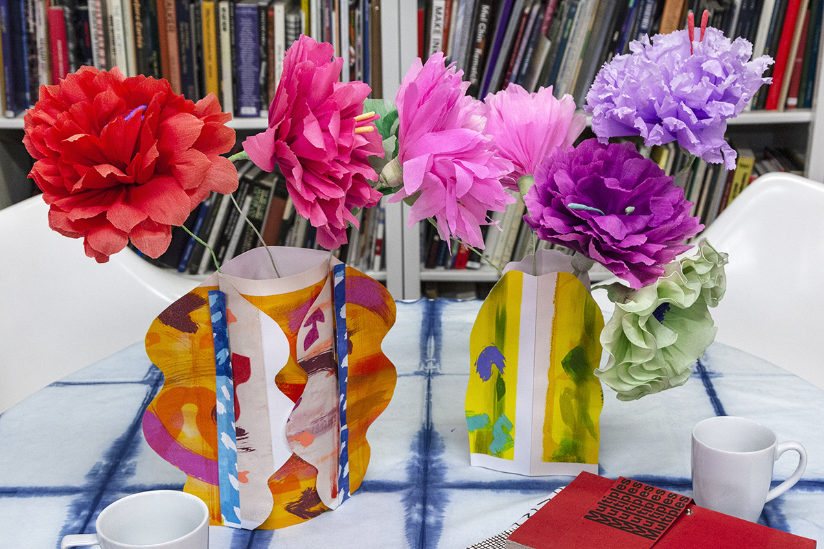 Two tabletop vases made of paper with colorful, abstract screenprinted designs holding bouquets of paper flowers.