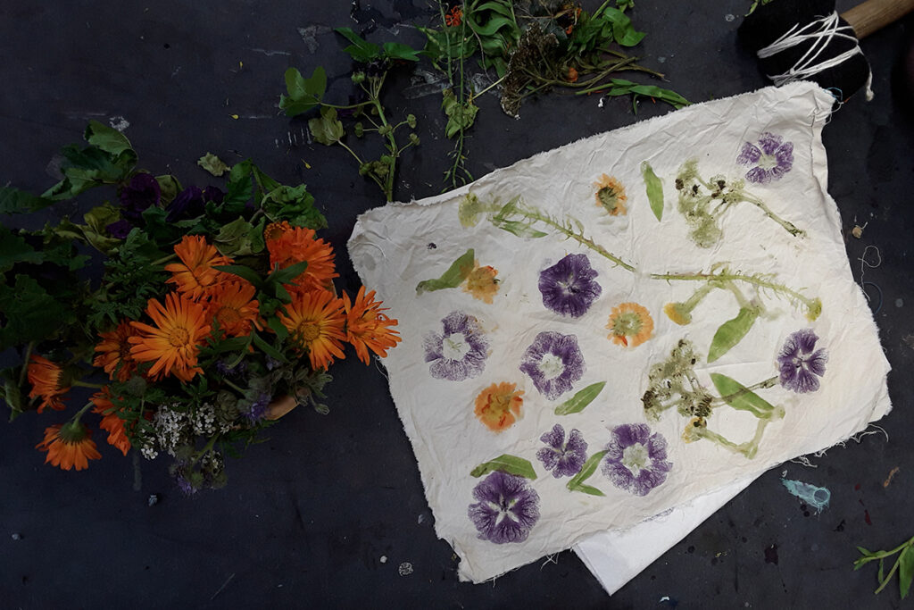 A cream colored napkin with prints of purple and gold flowers and green leaves and stems lies upon a surface near specimens of orange flowers.