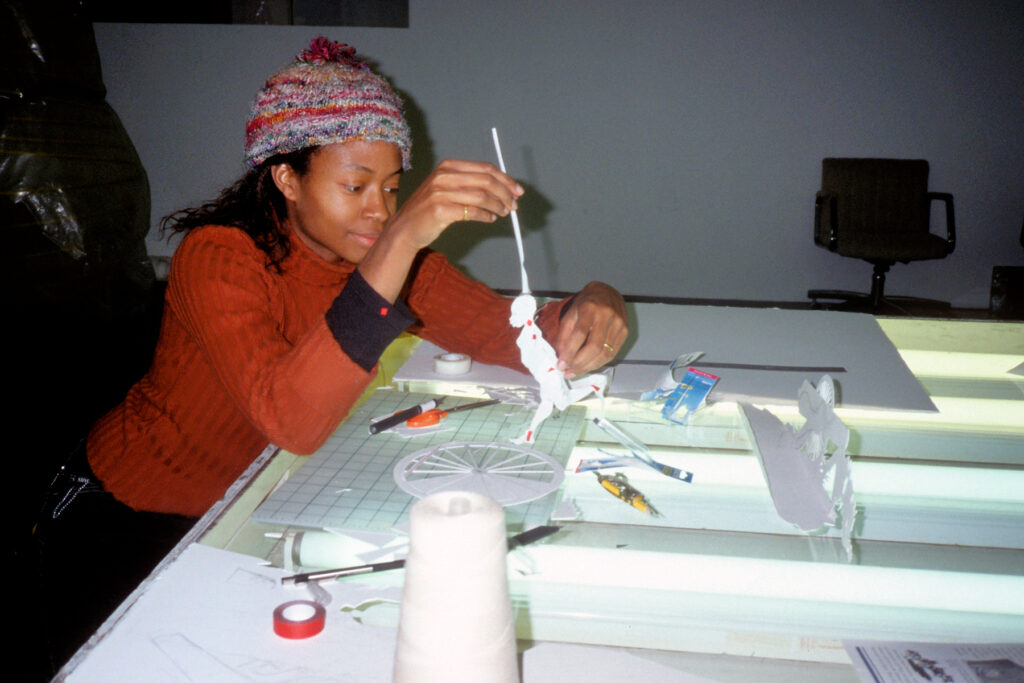 A photograph of Kara Walker, a young Black woman wearing an orange sweater and knitted cap, seated at a studio table while working with paper materials and tools.