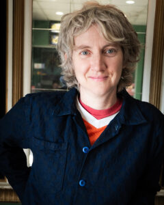 Portrait of Sharon Hayes, a middle-aged white woman with wavy, graying blonde hair. She is wearing a dark cardigan over a red and white collared t-shirt.