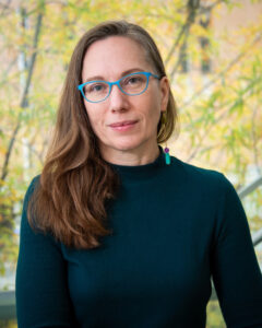 A portrait of Megan Voeller, a white person with long light brown hair and blue glasses. They are wearing a dark, long-sleeve shirt.