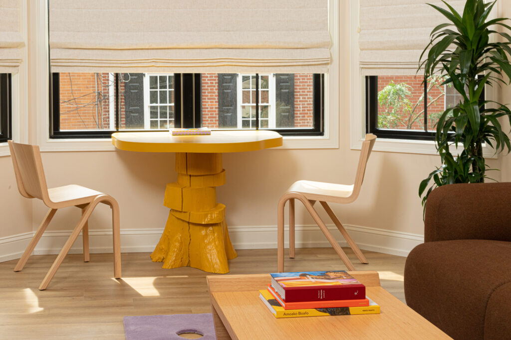 A photo of an interior space with two chairs and a small round table against a bay window with shades down. A potted plant and a coffee table are seen nearby.