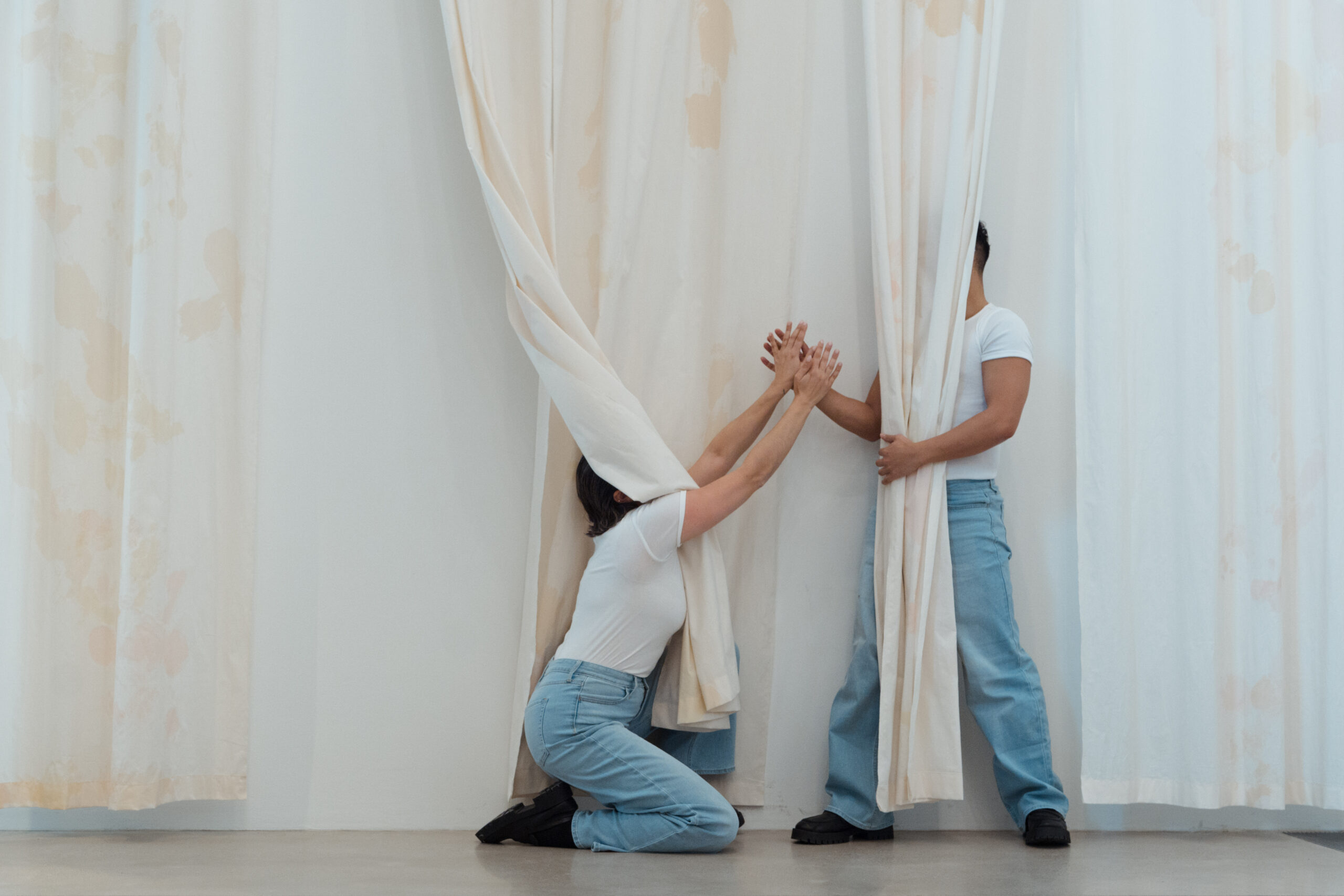 A photo of a performance with dancers' faces hidden behind a curtain, their hands raised upwards to reach one another. They are wearing white t-shirts and light blue jeans.