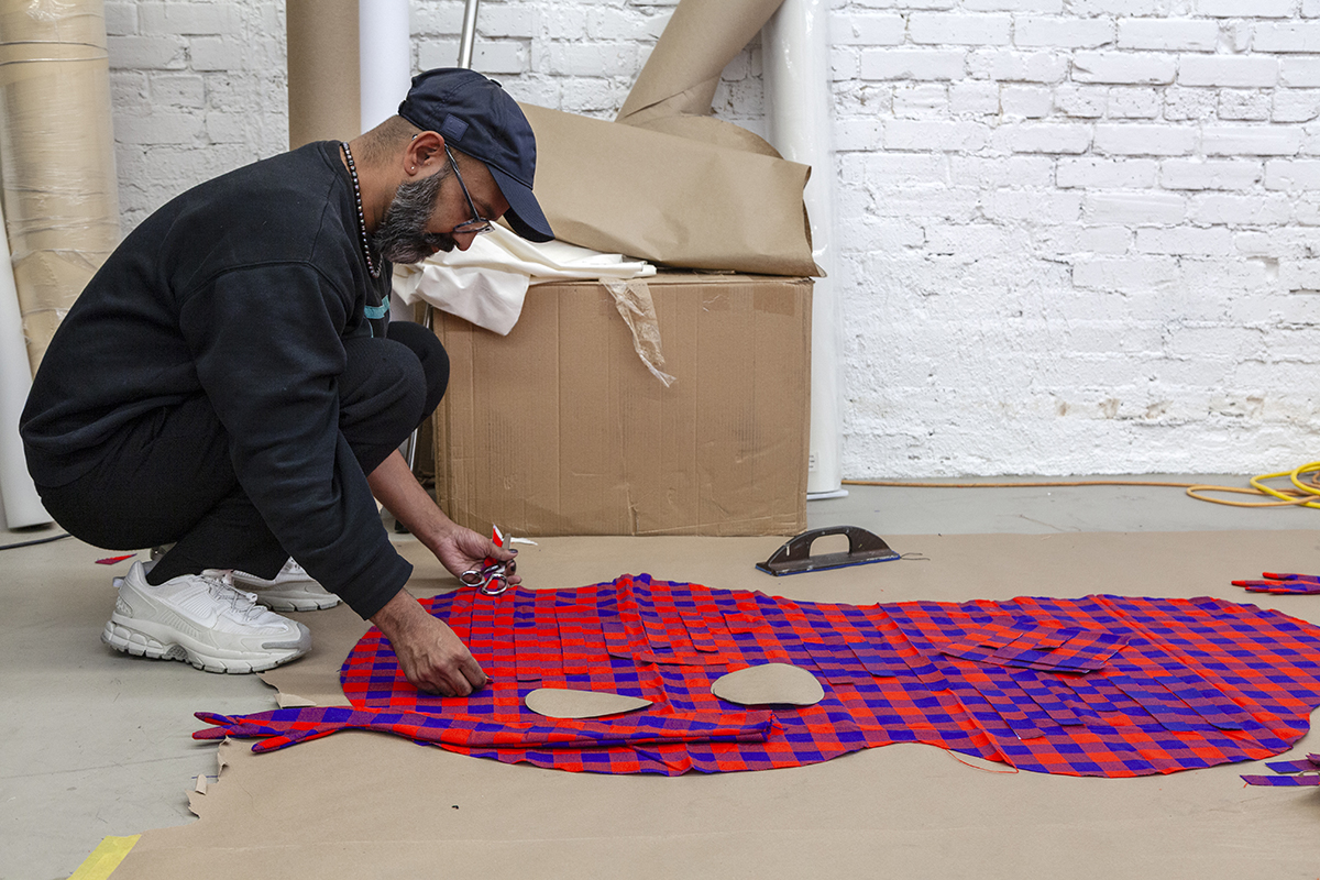 A photograph of an artist, Brendan Fernandes, squatting on the floor while working on an organically-shaped piece of plaid fabric. The artist is wearing all black, including a cap, long-sleeve t-shirt, and pants, with white shoes.